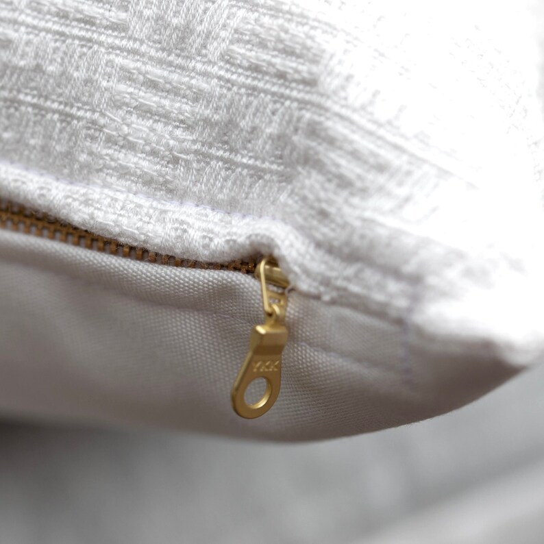 May include: Close-up of a white textured pillow with a gold zipper. The pillow has a woven pattern and a visible zipper pull. The zipper is partially open, revealing the pillow's interior.