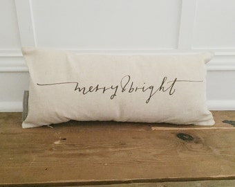 Puede incluir: Una almohada rectangular blanca con el texto "merry & bright" impreso en letra cursiva marrón.