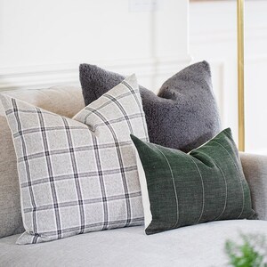 May include: Four decorative throw pillows in varying textures and patterns. One pillow features a gray plaid design, another is a solid gray, fluffy texture, and a third is a dark green with vertical white stripes. The pillows are arranged on a light gray sofa.