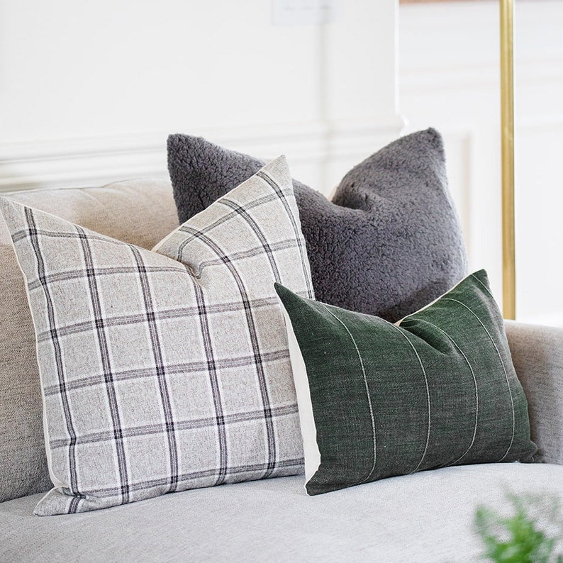 May include: Four decorative throw pillows in varying textures and patterns. One pillow features a gray plaid design, another is a solid gray, fluffy texture, and a third is a dark green with vertical white stripes. The pillows are arranged on a light gray sofa.