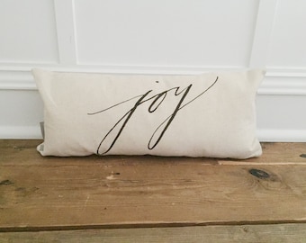 May include: A rectangular, off-white pillow with the word "joy" in elegant black script. The pillow is resting on a rustic wooden surface, with a white wall in the background.