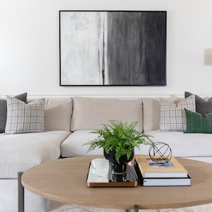 May include: A living room scene featuring a light-colored sectional sofa with decorative pillows in gray and plaid patterns. A round wooden coffee table holds books, a plant, and a decorative metal sphere. A large abstract painting hangs above the sofa.
