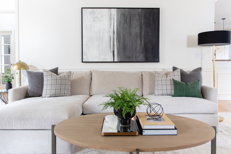 May include: A living room scene featuring a light-colored sectional sofa with decorative pillows in gray and plaid patterns. A round wooden coffee table holds books, a plant, and a decorative metal sphere. A large abstract painting hangs above the sofa.