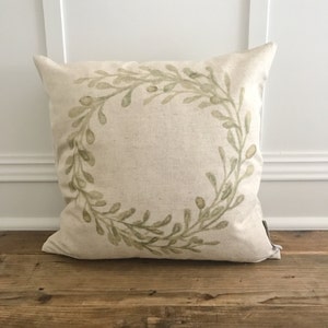 May include: A cream-coloured pillow with a green leafy wreath design. The pillow is made of a soft, natural fabric and has a subtle, rustic feel.