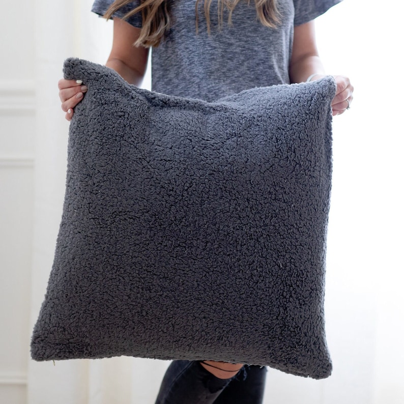 May include: A large, square, charcoal-colored pillow with a textured, plush surface. The pillow is held up by a person. The pillow appears to be made of a soft, fuzzy material, perfect for adding comfort and style to any living space.