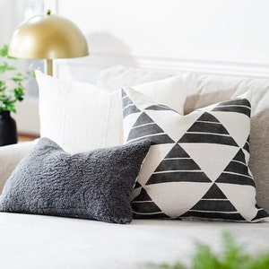 May include: A collection of decorative throw pillows in various textures and patterns. Includes a gray, fluffy pillow, a white textured pillow, and a pillow with a black and white geometric triangle design. The pillows are arranged on a neutral-colored sofa.