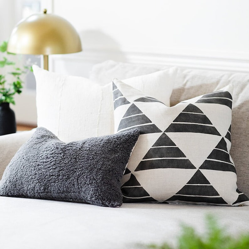 May include: A collection of decorative throw pillows in various textures and patterns. Includes a gray, fluffy pillow, a white textured pillow, and a pillow with a black and white geometric triangle design. The pillows are arranged on a neutral-colored sofa.