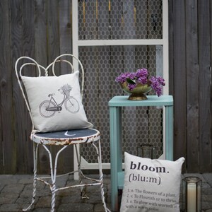 May include: A white metal chair with a floral patterned pillow and a bicycle design. A small teal table with a white pillow with the word "bloom" and its definition. A bouquet of purple flowers in a gold bowl sits on the table.