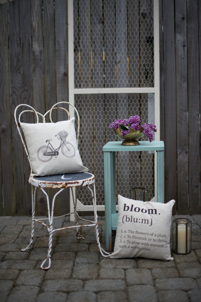 May include: A white metal chair with a floral patterned pillow and a bicycle design. A small teal table with a white pillow with the word "bloom" and its definition. A bouquet of purple flowers in a gold bowl sits on the table.