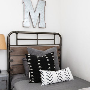 May include: A bed with a metal frame and a wooden headboard. The bed is made with gray bedding and several decorative pillows in shades of gray, black, and white. A large metal letter "M" hangs on the wall above the bed.