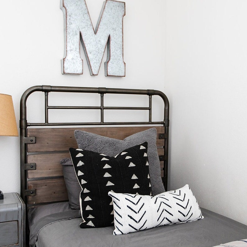 May include: A bed with a metal frame and a wooden headboard. The bed is made with gray bedding and several decorative pillows in shades of gray, black, and white. A large metal letter "M" hangs on the wall above the bed.