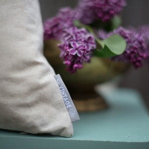 May include: A white pillow with a "Co. Vintage Chic" label on the side. The pillow is sitting on a light blue surface with a bouquet of purple lilac flowers in the background.