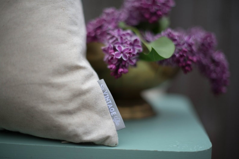 May include: A white pillow with a "Co. Vintage Chic" label on the side. The pillow is sitting on a light blue surface with a bouquet of purple lilac flowers in the background.