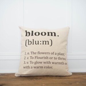 May include: A beige throw pillow with the word "bloom" and its phonetic spelling in black text. The pillow also includes a definition of the word "bloom" with three bullet points.