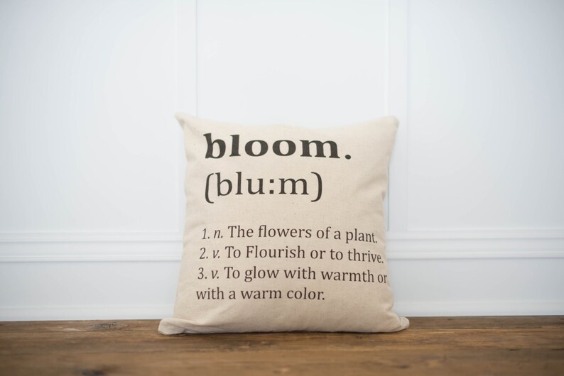 May include: A beige throw pillow with the word "bloom" and its phonetic spelling in black text. The pillow also includes a definition of the word "bloom" with three bullet points.