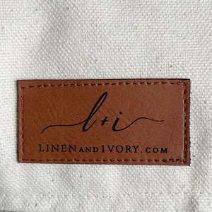 May include: A rectangular brown leather tag with the text "l + i" in a cursive font and "LINENANDIVORY.COM" in a sans-serif font. The tag is stitched around the edges and is set against a cream-colored fabric.