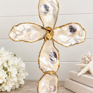 May include: A decorative cross sculpture constructed from five individual oyster shells. Each shell features a natural iridescent white interior and is edged with a metallic gold finish. The shells are arranged in a vertical cross formation.