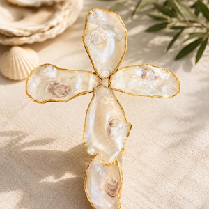 May include: A cross-shaped wall hanging constructed from five iridescent oyster shells. Each individual shell features a natural pearlescent finish and is outlined with a metallic gold-colored trim along the irregular, organic edges of the shell.