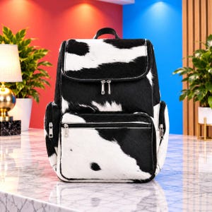May include: A backpack featuring a black and white cow print pattern with a textured hair-on-hide appearance. The bag includes a top flap, a front zippered pocket, and two side zippered pockets, all equipped with silver-toned metal hardware.