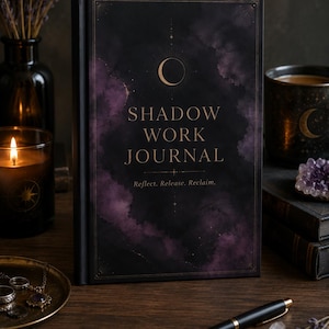 May include: A black hardcover journal with a purple and black nebula-like design on the front cover. The text reads SHADOW WORK JOURNAL and Reflect. Release. Reclaim. A gold crescent moon icon is centered above the title on the front cover.