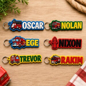 May include: Six rectangular keychains featuring colorful fire truck or fire hydrant graphics and the names Oscar, Nolan, Ege, Nixon, Trevor, and Rakim in bold lettering. Each keychain includes a silver metal ring for attachment.