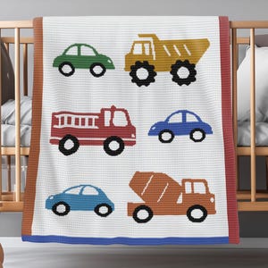 Könnte beinhalten: Eine weiße Babydecke mit bunten, gestickten Fahrzeugen, darunter ein grünes Auto, ein gelber Muldenkipper, ein rotes Feuerwehrauto, ein blaues Auto und ein orangefarbener Betonmischer. Die Decke hat braune, rote und blaue Ränder. Sie liegt auf einem Holzbettchen.