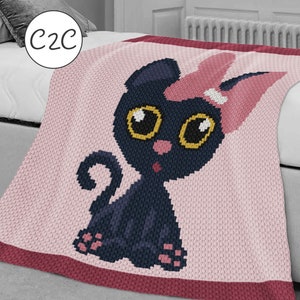 May include: A crocheted blanket featuring a black cat with pink ears and a pink bow. The cat has large yellow eyes and a pink nose. The blanket is pink with a burgundy border.