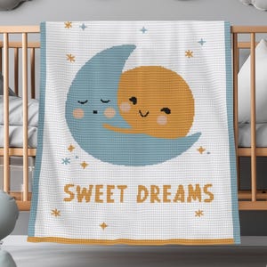 May include: A white baby blanket with a blue crescent moon and a yellow sun hugging each other. The blanket has the words "SWEET DREAMS" in gold letters, with stars and a gold border. The blanket is displayed in a wooden crib.