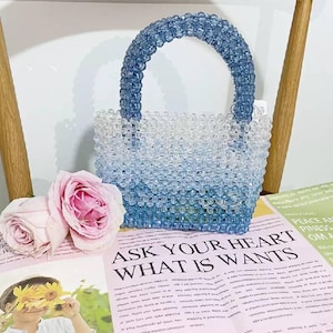 May include: A small handbag constructed from translucent blue and clear beads arranged in a gradient pattern. The bag features a rounded handle made of matching blue beads and a structured rectangular body. The beads are faceted and reflective.