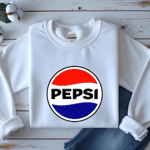 May include: A white long-sleeve crewneck sweatshirt featuring a circular Pepsi logo on the chest. The logo consists of a red upper section, a white middle wave, and a blue lower section, with the word PEPSI printed in bold black capital letters.