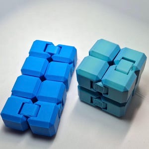 May include: Two geometric fidget toys made of interconnected plastic blocks. One toy is a solid bright blue rectangular shape, while the other is a square shape featuring a gradient of blue and teal tones with visible internal gear mechanisms.