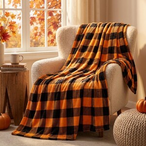 Peut inclure: Un plaid doux et moelleux orné d'un motif à carreaux vichy orange et noir. La couverture est drapée sur un fauteuil texturé de couleur crème, mettant en valeur son tissu épais et duveteux ainsi que son grand motif à carreaux répétitif.