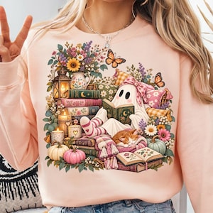 Peut inclure: Un sweat-shirt pêche clair avec une impression graphique représentant un fantôme blanc portant un nœud rose, lisant un livre entouré de piles de livres, d'un chat orange endormi, de citrouilles, de bougies, d'une lanterne, de fleurs et de papillons.