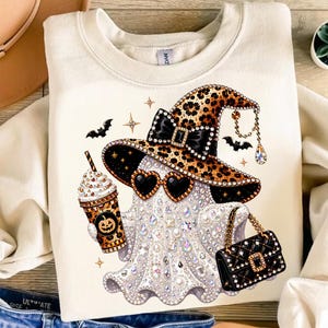 May include: A cream-colored crewneck sweatshirt featuring a graphic of a ghost wearing a leopard print witch hat with a black bow, heart-shaped sunglasses, and holding a leopard print coffee cup with a pumpkin icon and a black quilted handbag.