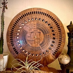 Puede incluir: Un calendario circular de madera con una mancha marrón oscura. El calendario presenta un dial circular con un dial interior más pequeño. El dial exterior muestra los días del mes y el año, mientras que el dial interior muestra los meses y el hemisferio. El calendario está marcado con los años 2020 a 2070.
