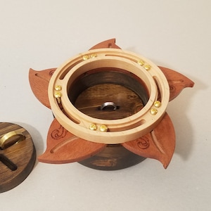 Puede incluir: Una caja de anillos de madera con diseño de flor. La caja tiene una base marrón oscuro, pétalos marrón rojizo y una parte superior de madera clara con detalles de latón. También se ve un soporte para anillos de madera separado.