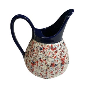 May include: A ceramic pitcher featuring a glossy dark blue handle and rim, contrasted by a textured, rounded body. The surface of the pitcher is finished with a speckled pattern of white, red, and dark blue flecks, creating a distinct visual depth.