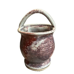 May include: A ceramic vessel with a rounded body, a flared base, and a single arched handle. The exterior features a mottled glaze with blended tones of deep reddish-brown, cream, and white, creating a textured appearance across the entire surface.