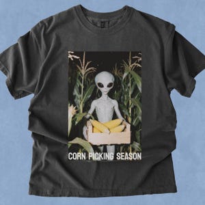 Op de afbeelding: Een antracietgrijs T-shirt met korte mouwen en een grafische print van een grijze alien in een maïsveld, die een houten kist met gele maïskolven vasthoudt. De tekst CORN PICKING SEASON staat in witte hoofdletters onder de kist gedrukt.