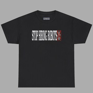 Könnte beinhalten: Ein schwarzes T-Shirt mit kurzen Ärmeln und dem weißen Schriftzug STOP HIRING ROBOTS auf der Brust. Neben dem Text befindet sich eine weiße Grafik eines Roboters in einem roten Rechteck mit einem roten X darüber. Das Shirt hat einen Rundhalsausschnitt.
