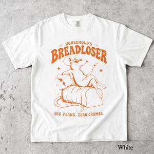 May include: A white t-shirt featuring an orange graphic of a mouse wearing sunglasses, lounging on a loaf of bread. The text reads HOUSEHOLD'S BREADLOSER above the mouse and BIG PLANS. ZERO CRUMBS. below the mouse, with small star accents.