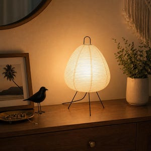 May include: A teardrop-shaped table lamp with a ribbed white paper shade and a thin black metal wire frame. The lamp features a small black handle at the top and stands on three slender black metal legs, emitting a warm, soft glow from within.