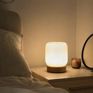 May include: A cylindrical table lamp with a white, vertically ribbed shade and a circular wooden base. The lamp emits a warm, soft glow, illuminating the surrounding area with a gentle light, creating a cozy atmosphere in the room.