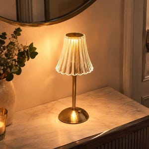 May include: A table lamp featuring a clear, fluted glass shade with a scalloped edge, supported by a slender gold-toned metal stem and a circular base. The lamp emits a warm, soft glow, highlighting the vertical ridges of the glass shade.