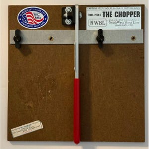 Op de afbeelding: Een gereedschap met een bruine houten basis, gelabeld als The Chopper Tool #49-4 van NorthWest Short Line. Het bevat een verticaal metalen snijmes met een rode handgreep, een horizontale metalen geleiderail met twee zwarte knoppen en waarschuwingen.