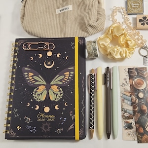 Peut inclure: Un agenda 2026-2027 avec une couverture papillon et lune, une trousse en velours côtelé beige marquée YUEBU, un chouchou jaune, quatre stylos, un rouleau de ruban adhésif, un porte-clés en perles, un tampon et un marque-page illustré de livres.