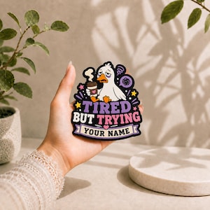 Può includere: Un adesivo fustellato raffigurante un'anatra bianca dei cartoni animati con gli occhi stanchi che tiene in mano una tazza di caffè. Il testo recita TIRED BUT TRYING e include uno spazio per YOUR NAME. Il design usa accenti viola, rosa e gialli su sfondo nero.