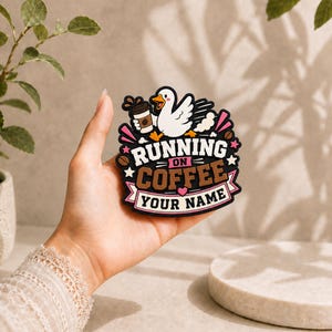 May include: A rubberised patch featuring a white cartoon goose holding a coffee cup, with the text RUNNING ON COFFEE and YOUR NAME in brown and white lettering, accented by pink stars, hearts, and brown coffee beans on a black background.