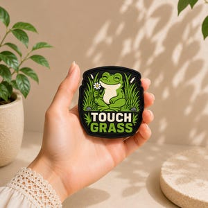 Puede incluir: Un parche de goma negro con una rana verde de dibujos animados sentada en hierba alta y sosteniendo una margarita blanca. El texto TOUCH GRASS está impreso en letras mayúsculas blancas y verdes en la parte inferior del parche.