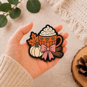 Op de afbeelding: Een rubberen patch met een oranje mok gevuld met slagroom, versierd met groene hartjes. Het ontwerp bevat een crèmekleurige pompoen, een groot oranje esdoornblad, een roze strik en twee bruine kaneelstokjes, allemaal omlijnd met zwart.